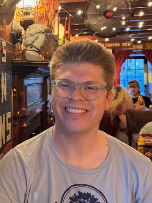 Smiling person with short hair and clear glasses sits in a warmly lit retro-style restaurant, with string lights and decor in the background.