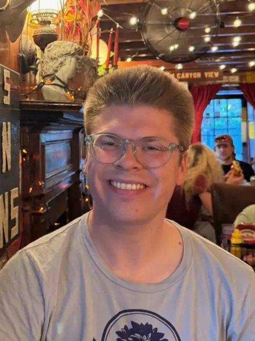 Smiling person with short hair and clear glasses sits in a warmly lit retro-style restaurant, with string lights and decor in the background.