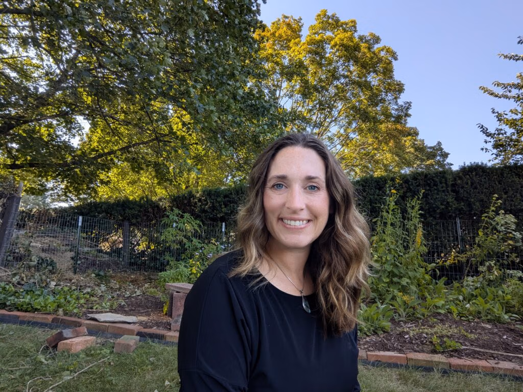 Smiling woman in a black top outdoors, with lush green trees, a wire fence, and garden plants in the background on a sunny day.