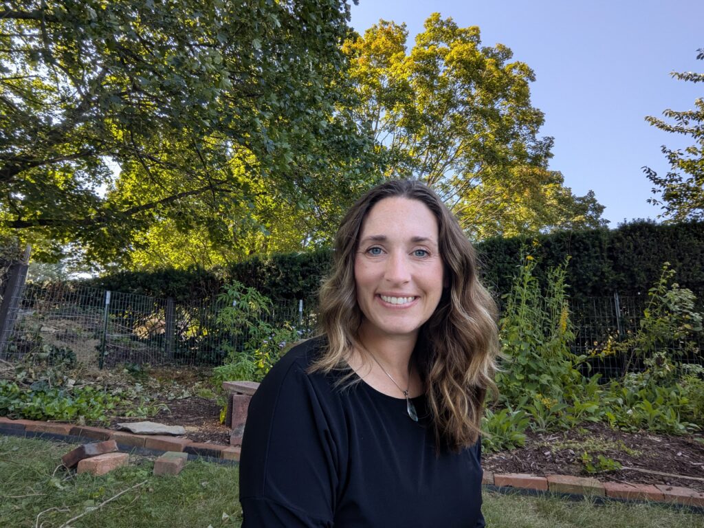 Smiling woman in a black top outdoors, with lush green trees, a wire fence, and garden plants in the background on a sunny day.