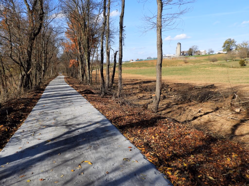 Tree-lined concrete path through a fall forest with leaves on the ground and a distant farm with a silo under a blue sky from the trail.