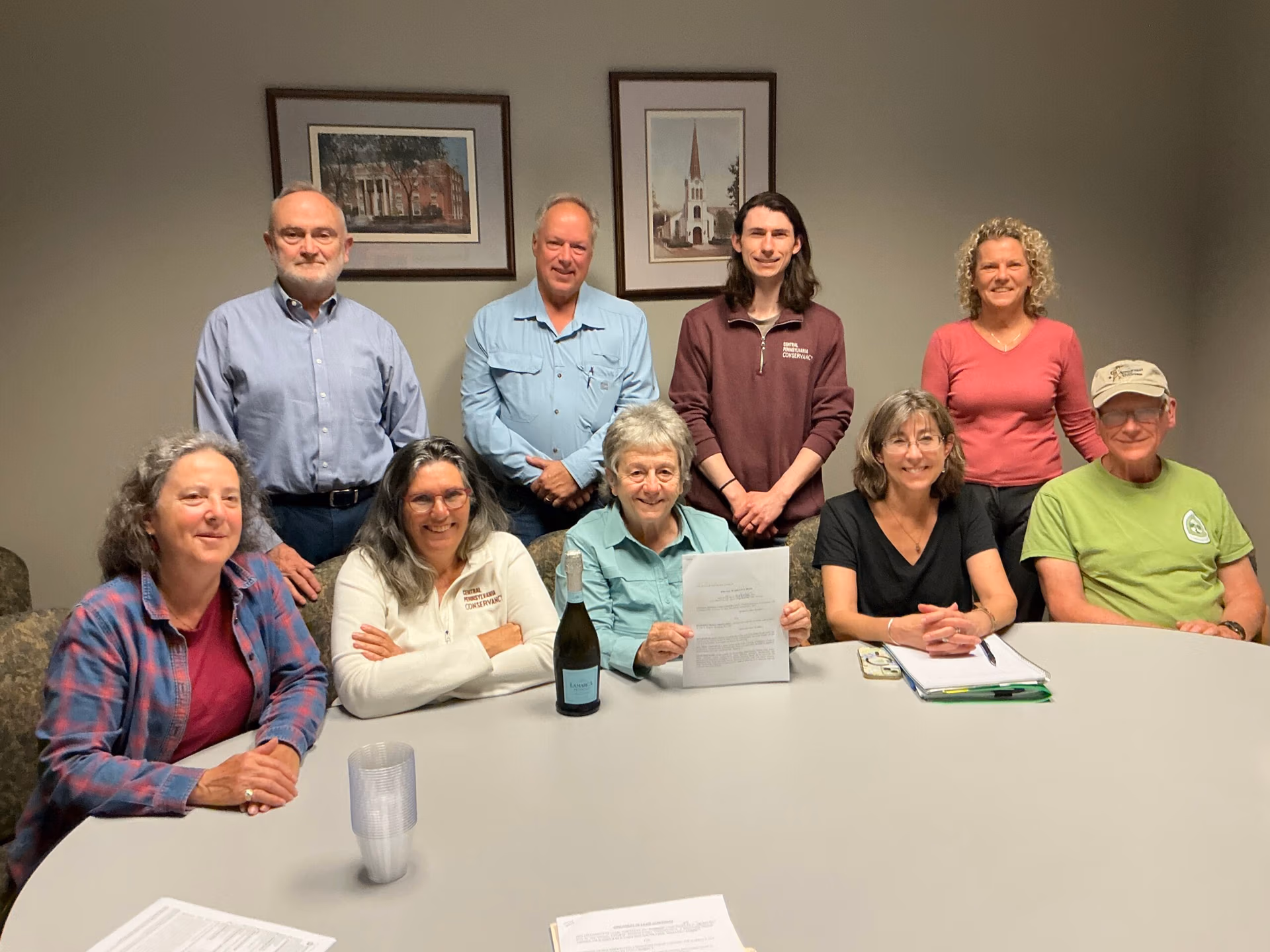 Group of nine adults posing for a photo in a meeting room, some standing behind a seated row of coworkers, papers and a wine bottle on the table.